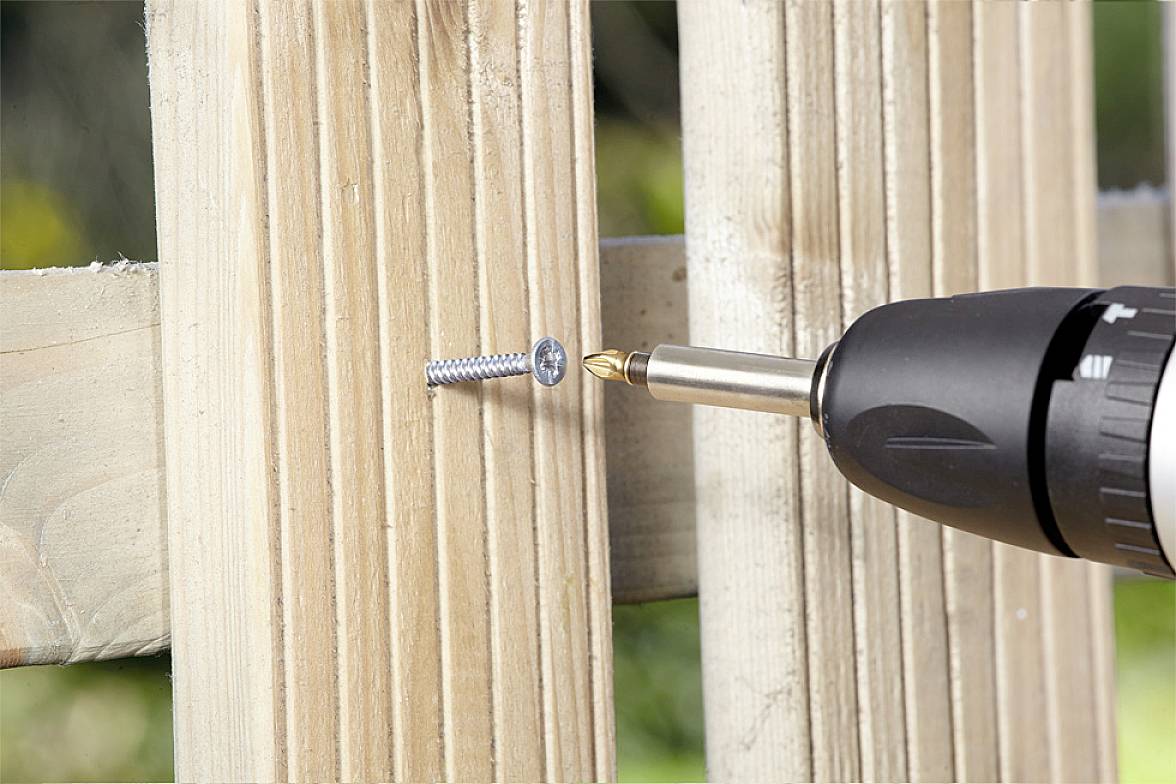 Cordless drill driving a screw into a wooden fence. Focus on the screw and drill bit, depicting DIY or construction work.