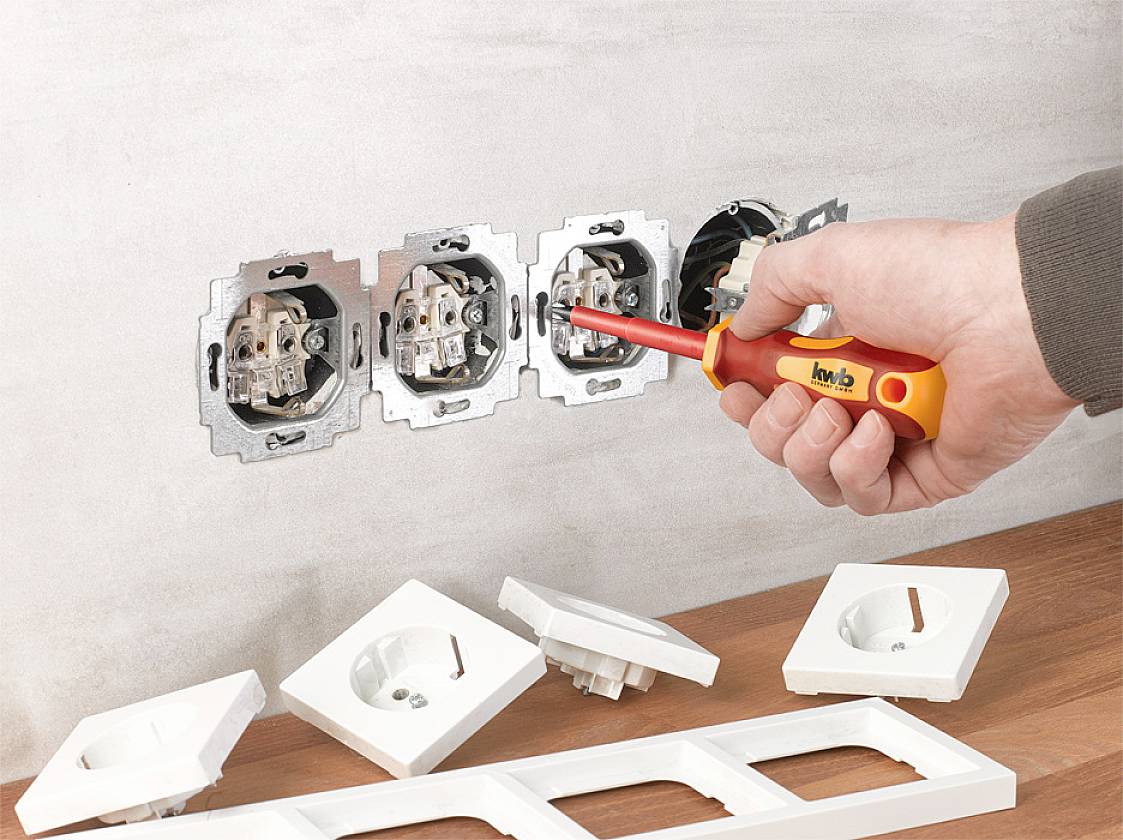 A hand with a screwdriver is installing sockets into the wall. Beside it, socket covers and socket plates are lying on the table.