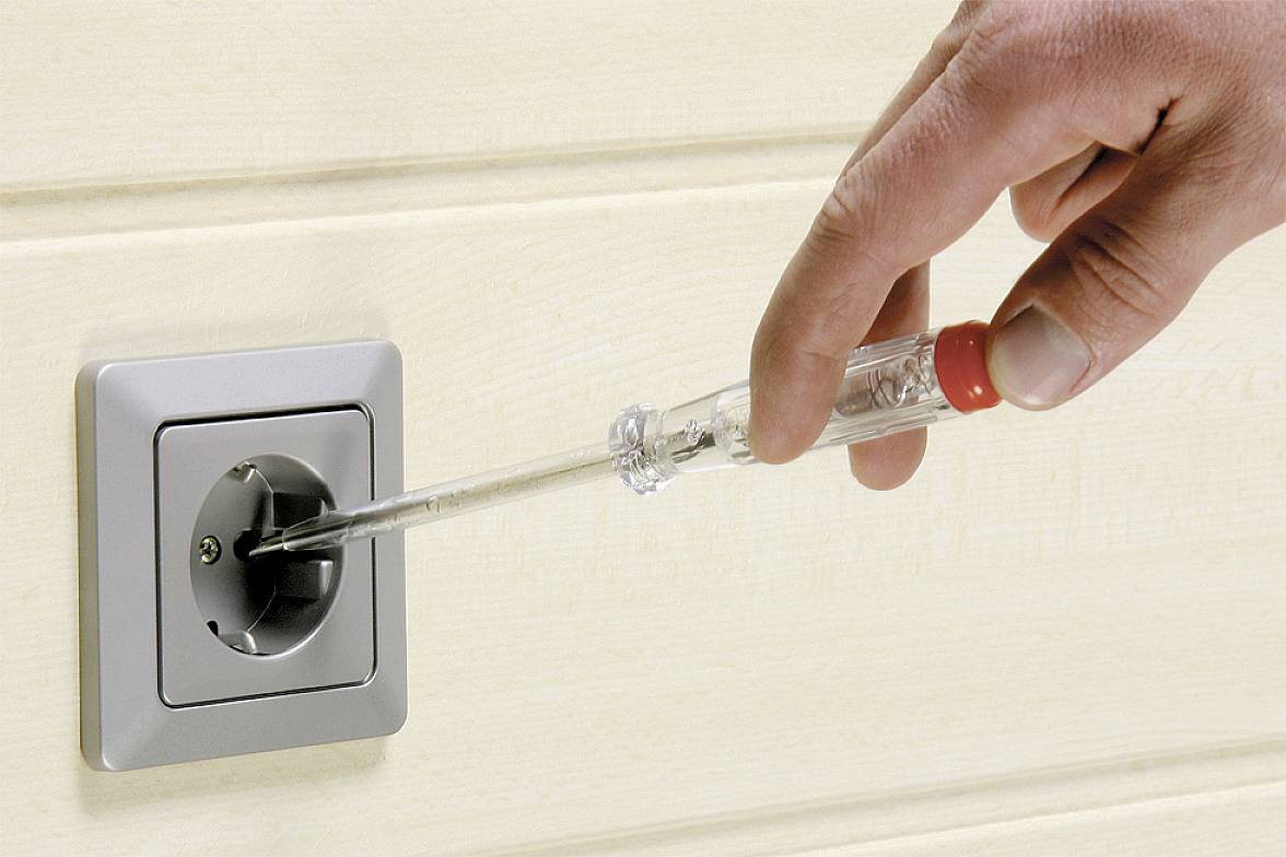 A hand holds a screwdriver and checks a socket on a wall.