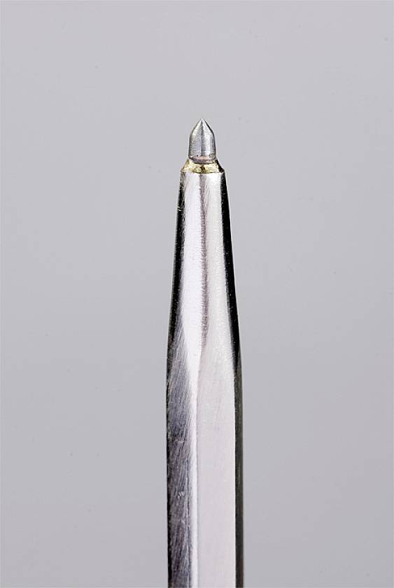 Close-up of a ballpoint pen nib on a grey background. The shiny metallic tip with the small ball at the end is visible.