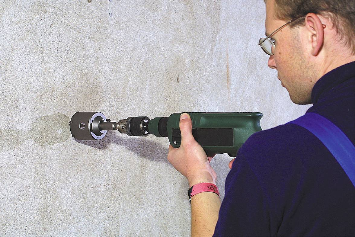 A man is drilling a hole in a concrete wall using an electric drill. He is wearing safety glasses and work clothing.