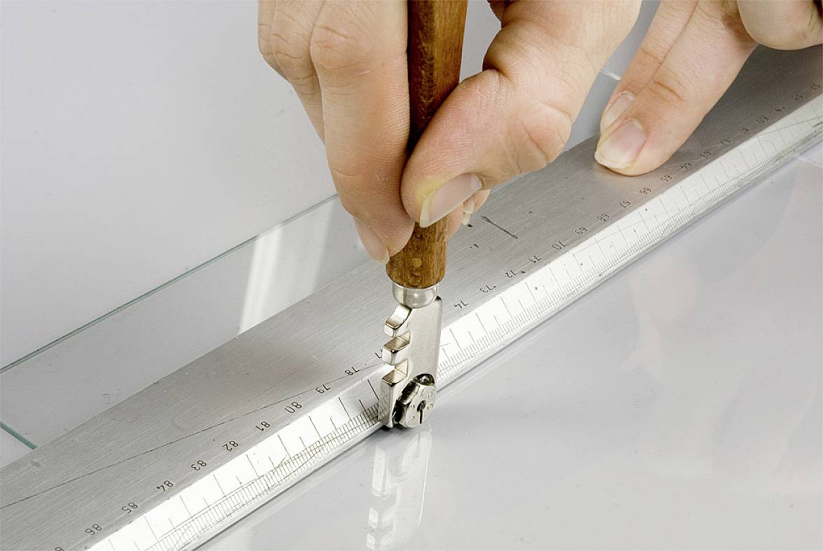 A hand is using a glass cutter with a wooden handle to cut along a metal ruler on a glass pane.