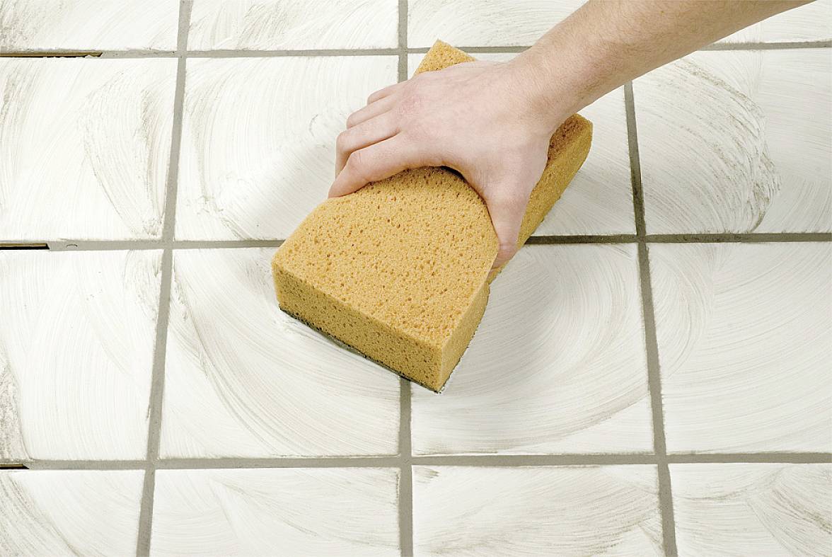 A hand cleans white tiles with a sponge, creating circular cleaning marks.