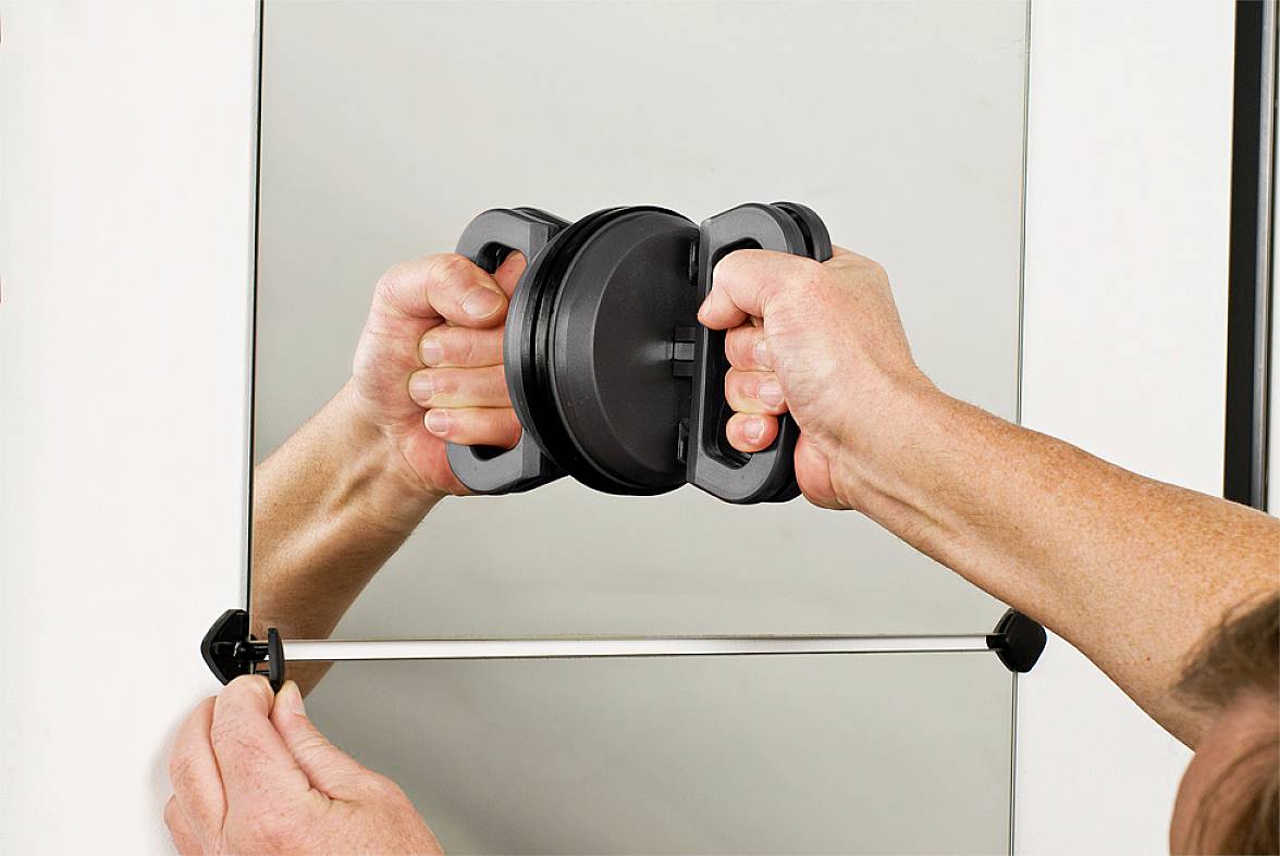 A person is holding a round suction cup handle against a glass pane. Hands are securing a metal clip at the bottom.