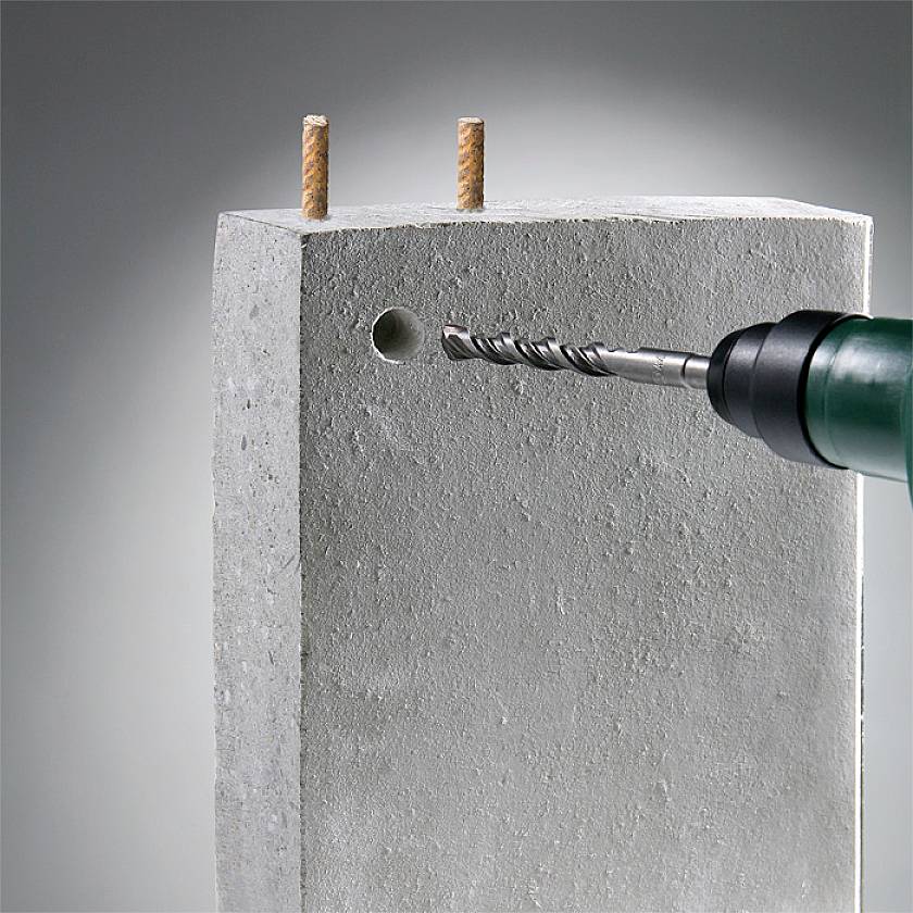 A drill is boring a hole in a concrete wall. Two wooden wall plugs are already inserted at the top of the wall.