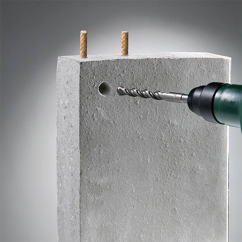 A drill is drilling a hole in a concrete block. Two wall plugs are already secured at the top of the concrete block.