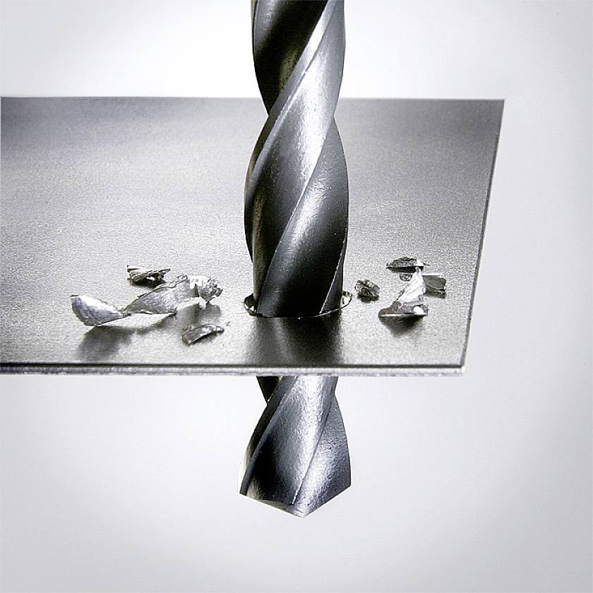 A metal drill pierces a metal plate, surrounded by metal shavings.