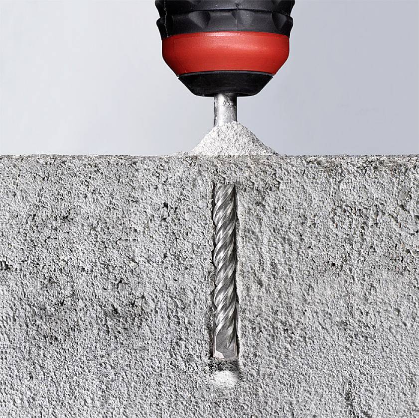 A drill with a black and red handle is drilling a hole into a grey concrete surface. The drill is generating dust as it penetrates.