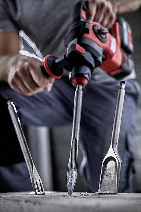 A person is holding a red rotary hammer drill with various attachments in front of them, ready for use on a concrete surface.