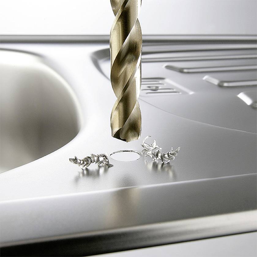 A drill penetrates stainless steel in a kitchen. Metal shavings surround the hole being drilled into a worktop.