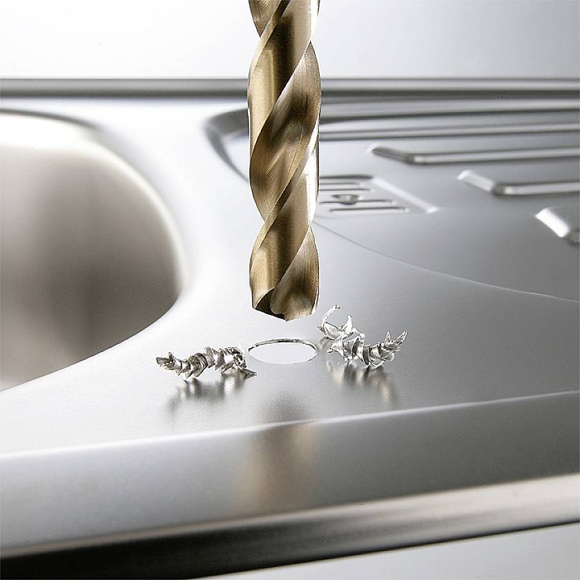 A drill is piercing a stainless steel sink. Metal shavings are scattered around the hole.