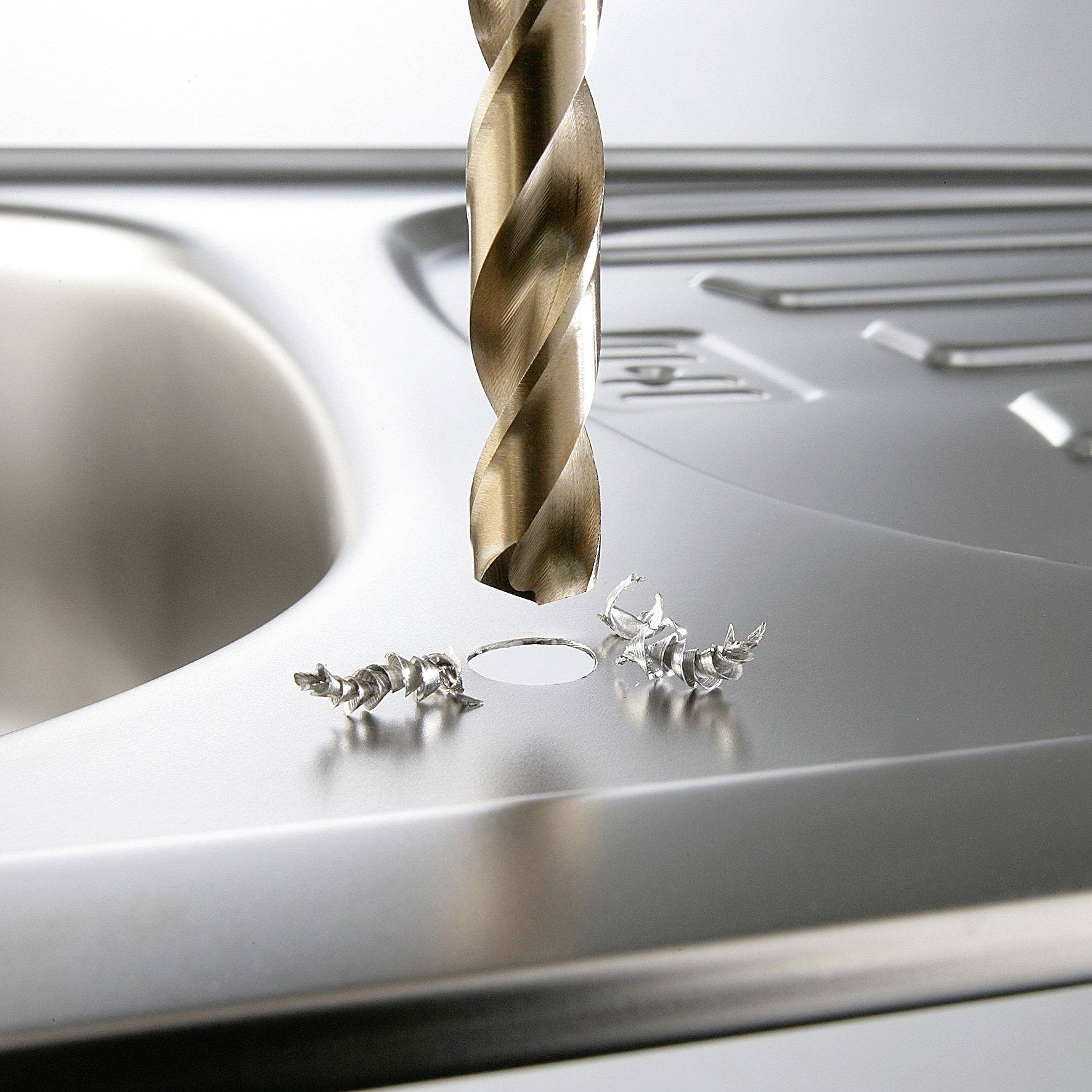 A metal drill penetrates a stainless steel sink, with spirals of metal shavings visible around the drill hole.