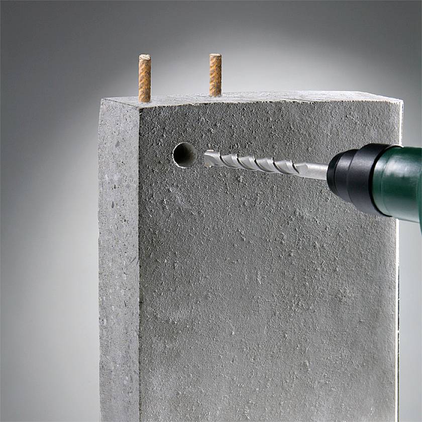 A concrete block is being drilled through by a drill bit. Two rods are fixed in the block, protruding from the top of the concrete.