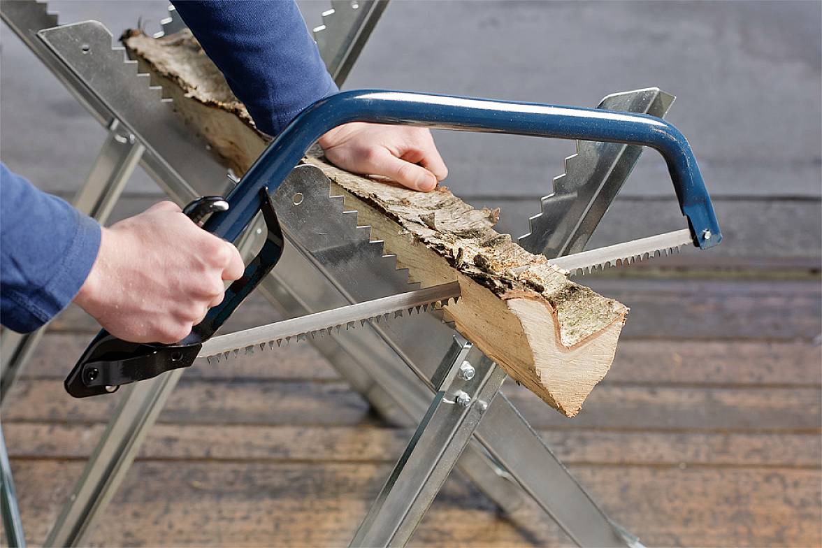A person is sawing a wooden log with a bow saw on a metal sawbuck stand.