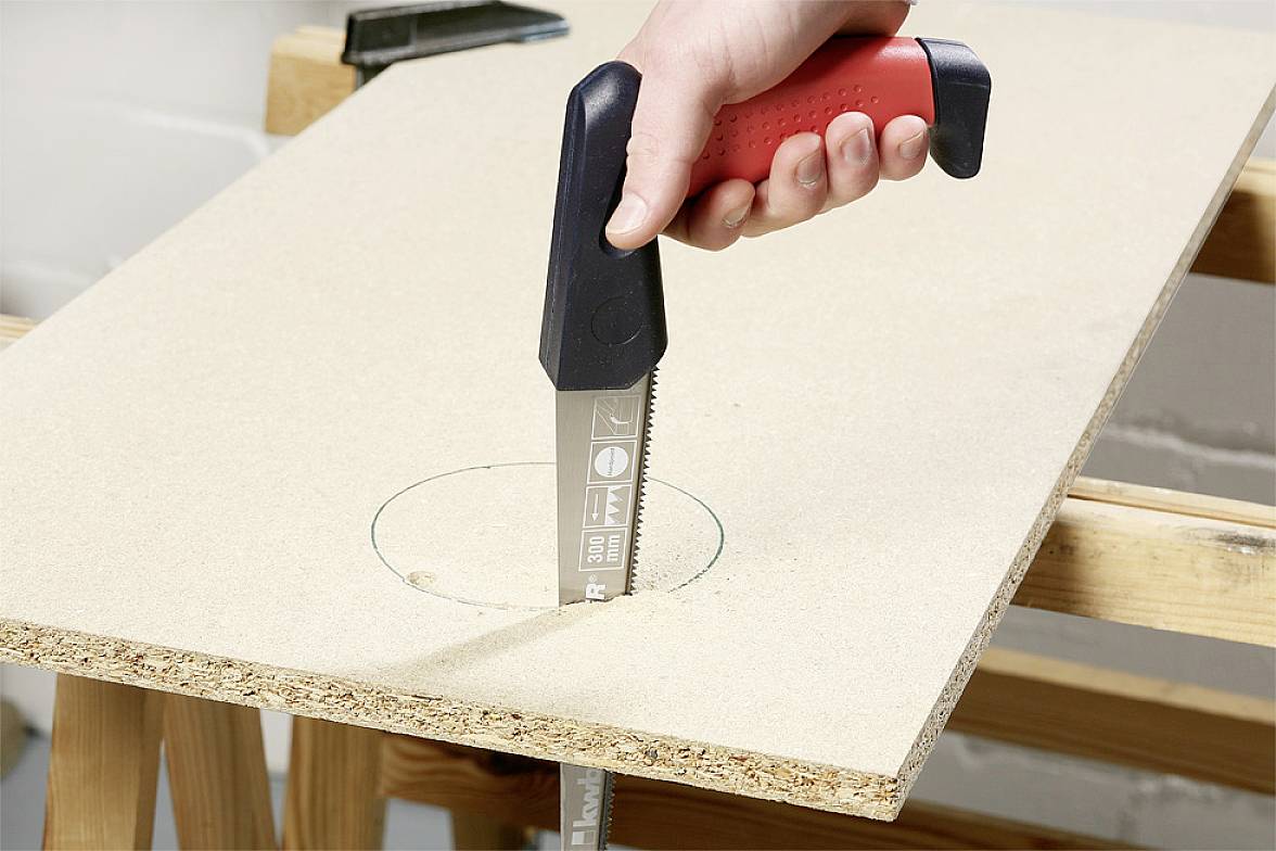 A hand is holding a red and black saw and cutting a circular hole in a chipboard panel on a workbench.