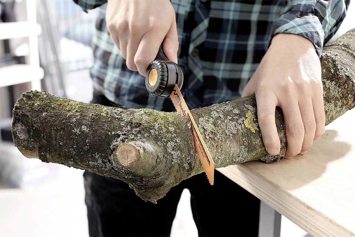A person in a checked shirt is cutting a thick branch with a handsaw on a workbench.