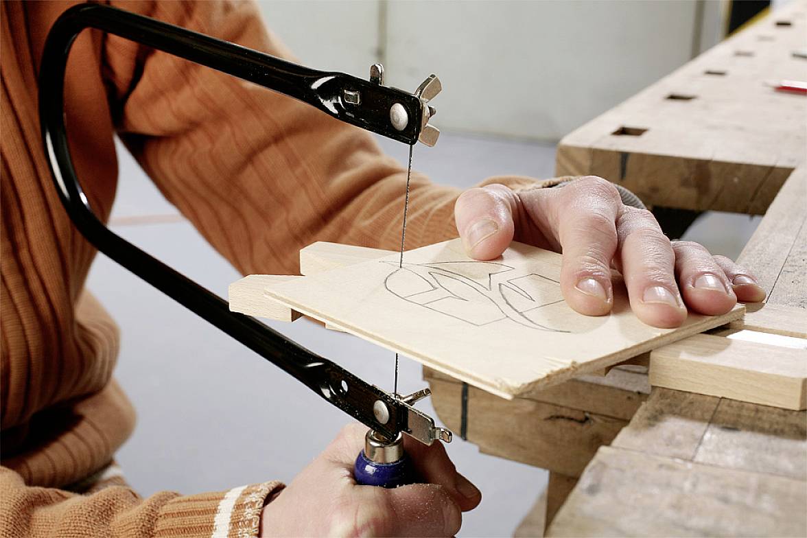 A person is cutting a pattern into a piece of wood using a fretsaw, which is secured to a workbench.