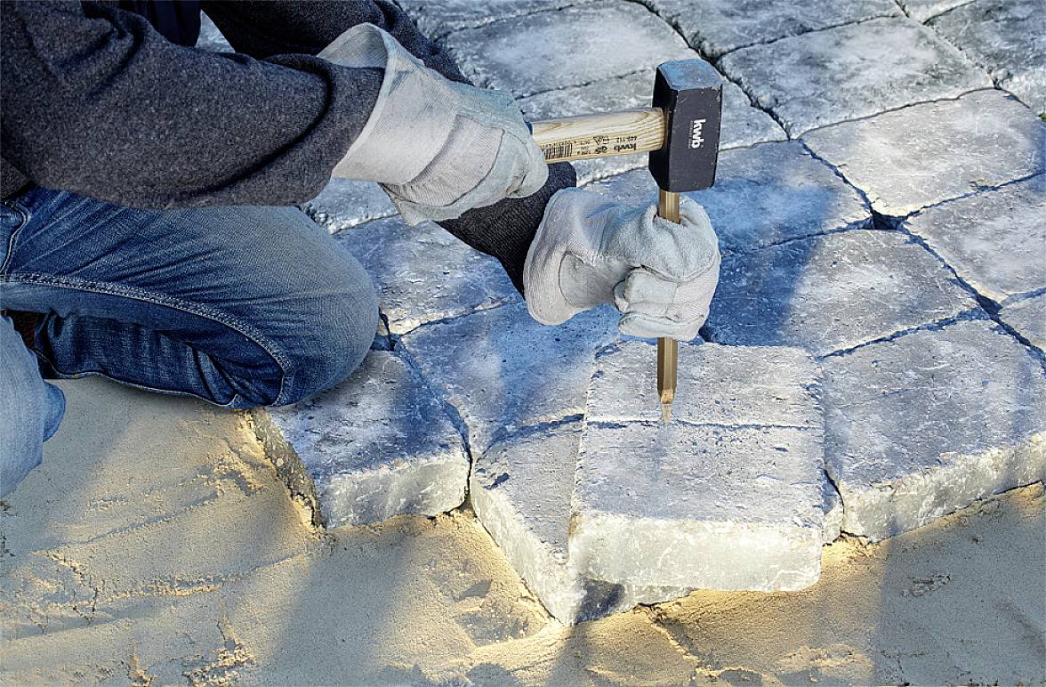 A person is laying cobblestones with a hammer and chisel on sandy ground. The hands are wearing protective gloves.