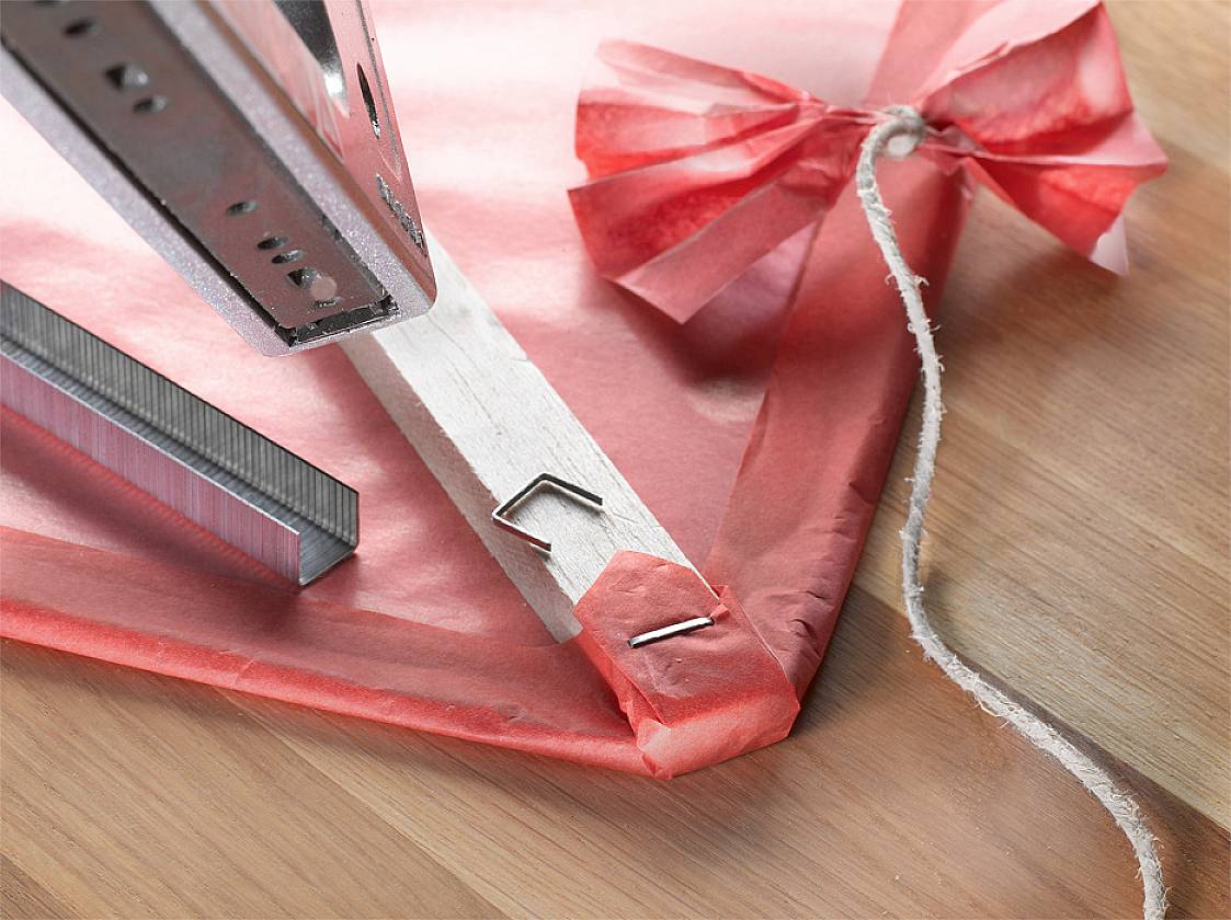 A red paper dragon, fastened to a wooden lath with a drawing pin. A piece of string lies on a wooden surface.