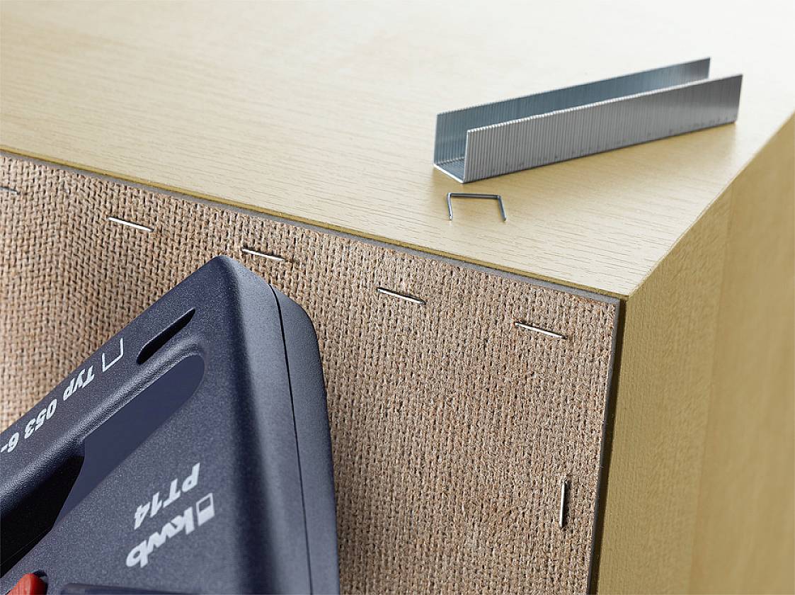 A piece of furniture is secured with a staple gun and staples. The staple gun is black, and staples are lying on the corner.