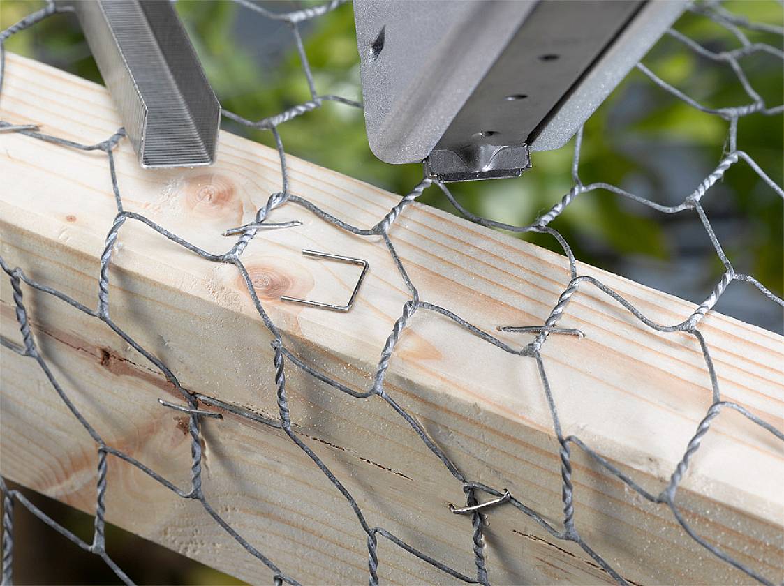 A piece of wire mesh is fastened to a wooden frame with metal staples. Green plants can be seen in the background.