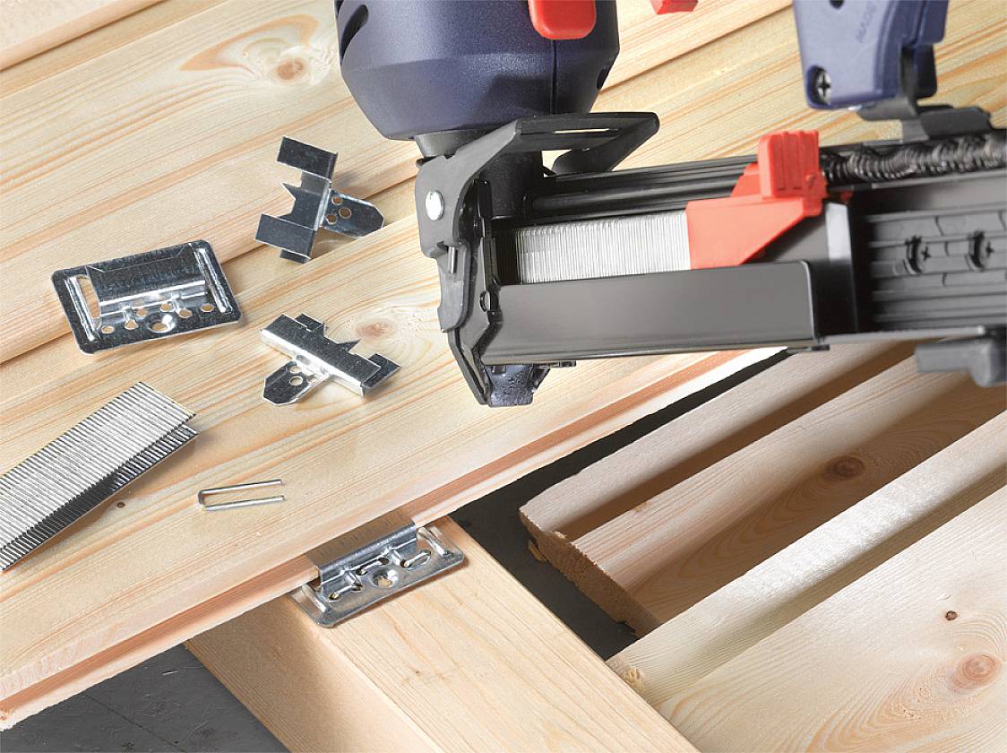 A nail gun secures wooden battens with metal fixings and nails. Various types of fittings and nails are laid out on the battens.