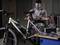 A man is working on a bicycle in a workshop, surrounded by tools and equipment on a table.