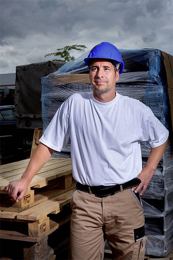 A man in workwear and a blue hard hat stands outside in front of stacked wooden pallets, gazing into the distance.