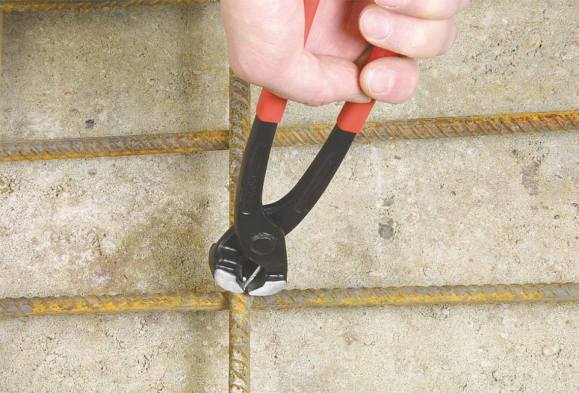 A hand is using bolt cutters to cut a rusty wire rope on a concrete surface.