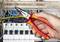 A hand is holding a pointed pliers and working on cables in an electrical cabinet, surrounded by coloured wires and circuit breakers.