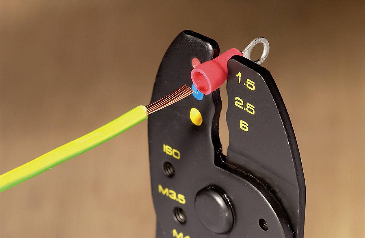 Wire strippers cut through a yellow cable. The pliers have markings for wire sizes. A red cable lug is attached to a wire.
