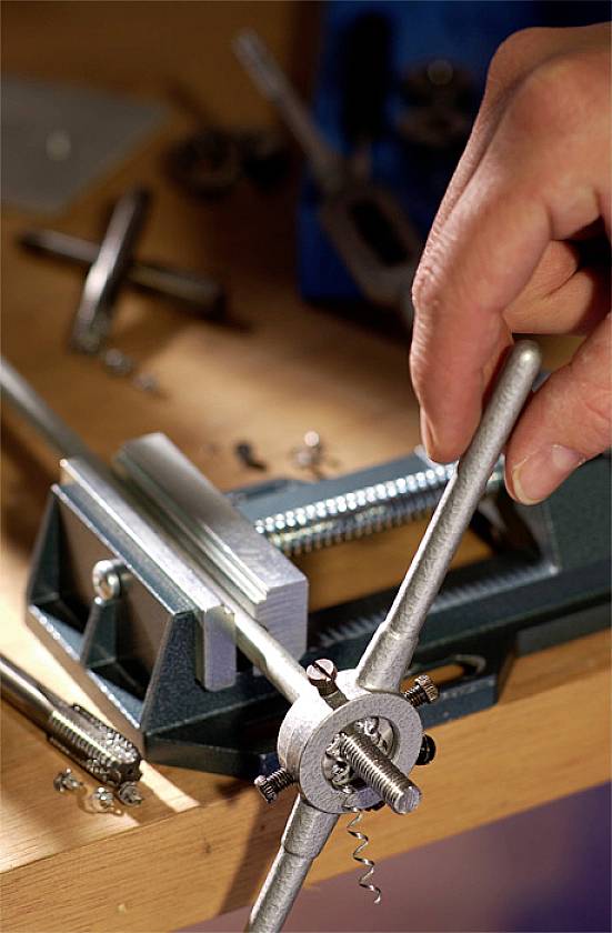 A hand is using a thread-cutting tool to cut a thread. A vice is holding the metal rod on a work surface.