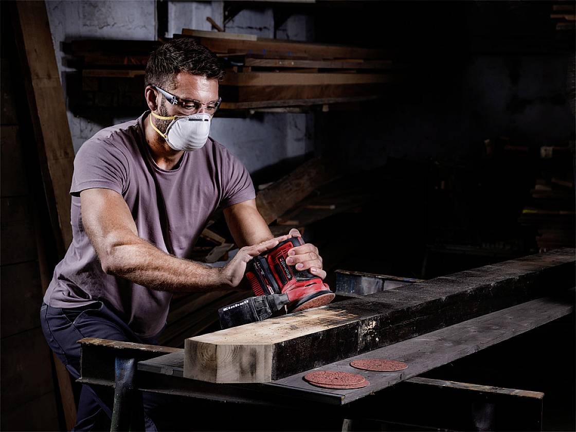 A man is sanding a wooden board in a workshop. He is wearing safety glasses and a mask to protect himself from dust.