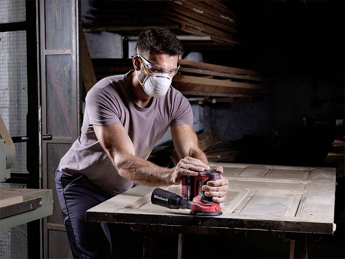 A man wearing safety glasses and a respiratory mask is grinding a wooden board with an electric sander in a workshop.