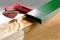 A green metal pipe lies next to a wooden beam that is being partially sanded. A red sanding device is positioned nearby.