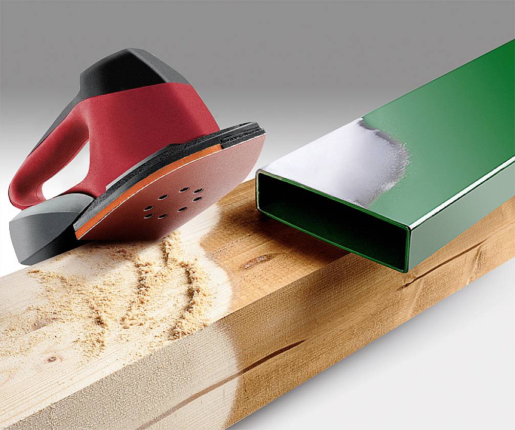 A red and black grinding tool smooths a piece of wood, with a shiny green metal profile visible on the right.