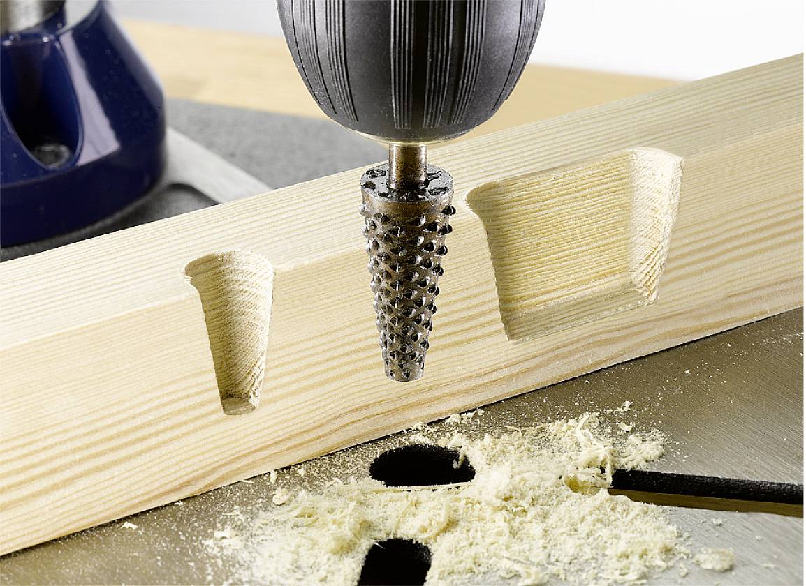 A wooden hole is created using a cone-shaped drill bit. To the right of the image, a completed hole in the wood is already visible. Wood shavings can be seen.