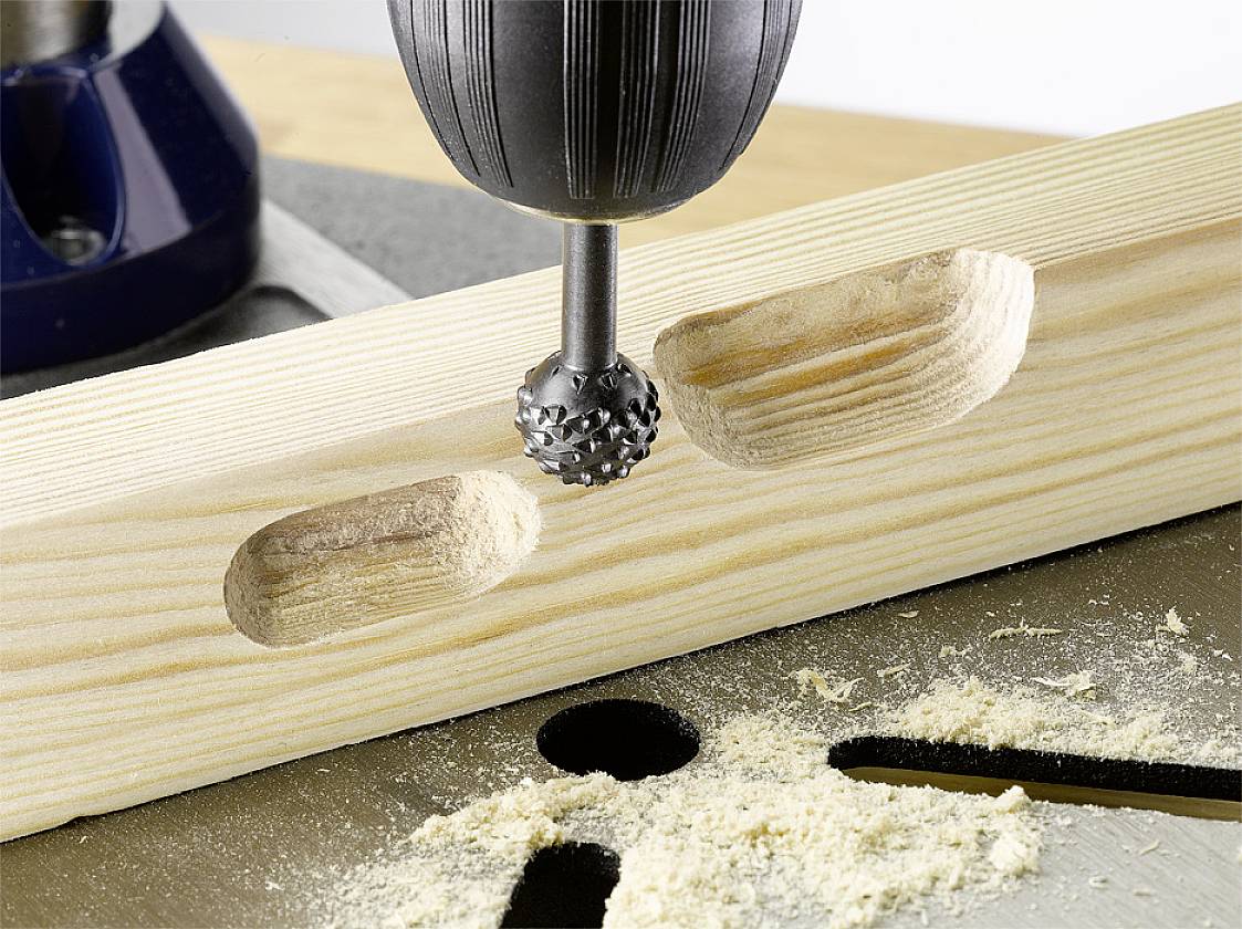 A drill is cutting oval recesses into a wooden board. Sawdust is scattered around the holes, with the table surface visible.