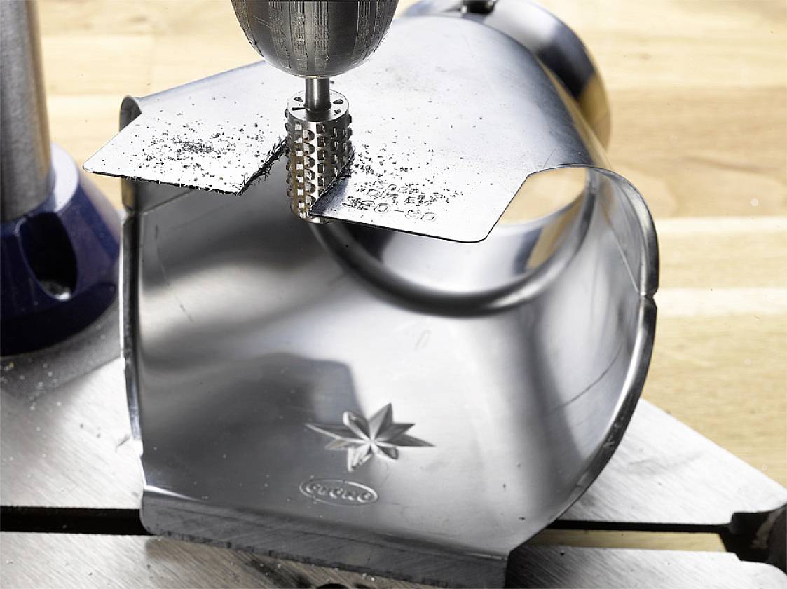 A metal drill machine is machining a curved metal piece. Metal shavings are visible. A wooden surface lies in the background.