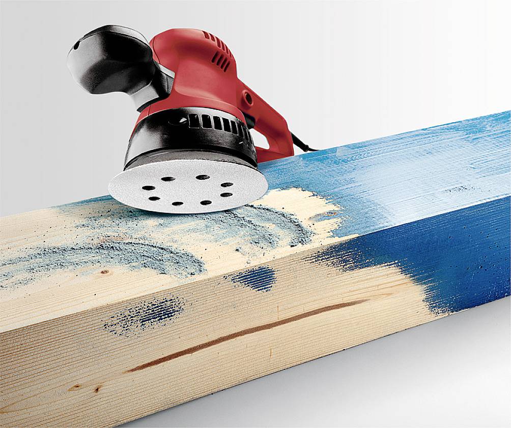 A red orbital sander is working on a wooden board. The sander is removing blue paint from the wood surface.