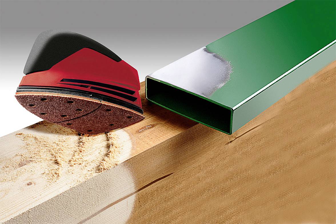 A red hand sander is working on wood, next to a green metal pipe with a shiny surface.