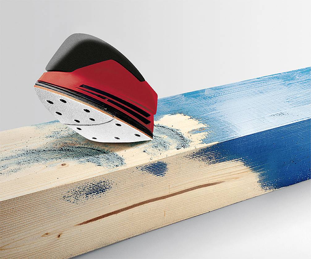 A red grinding tool sands a wooden surface that is partially painted blue. Dust is created by the movement of the tool.