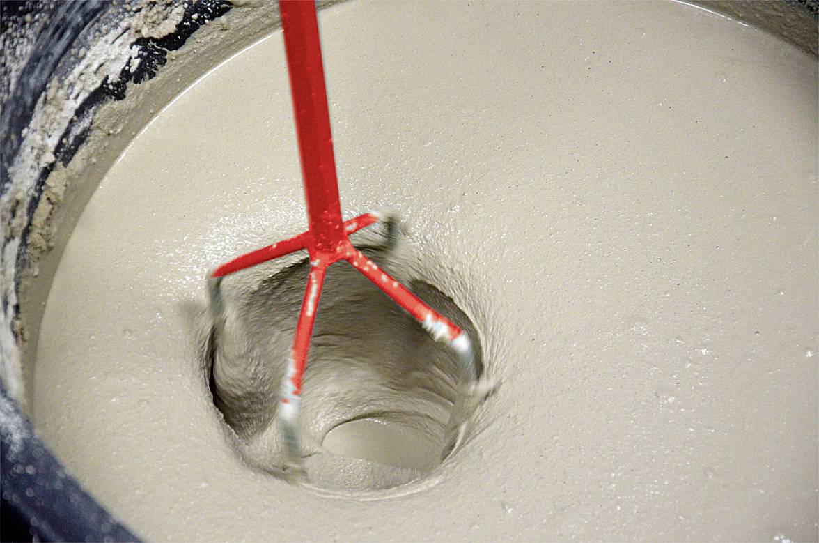 A rotating mixing rod stirs grey concrete in a bucket. The concrete is liquid and is thoroughly mixed.