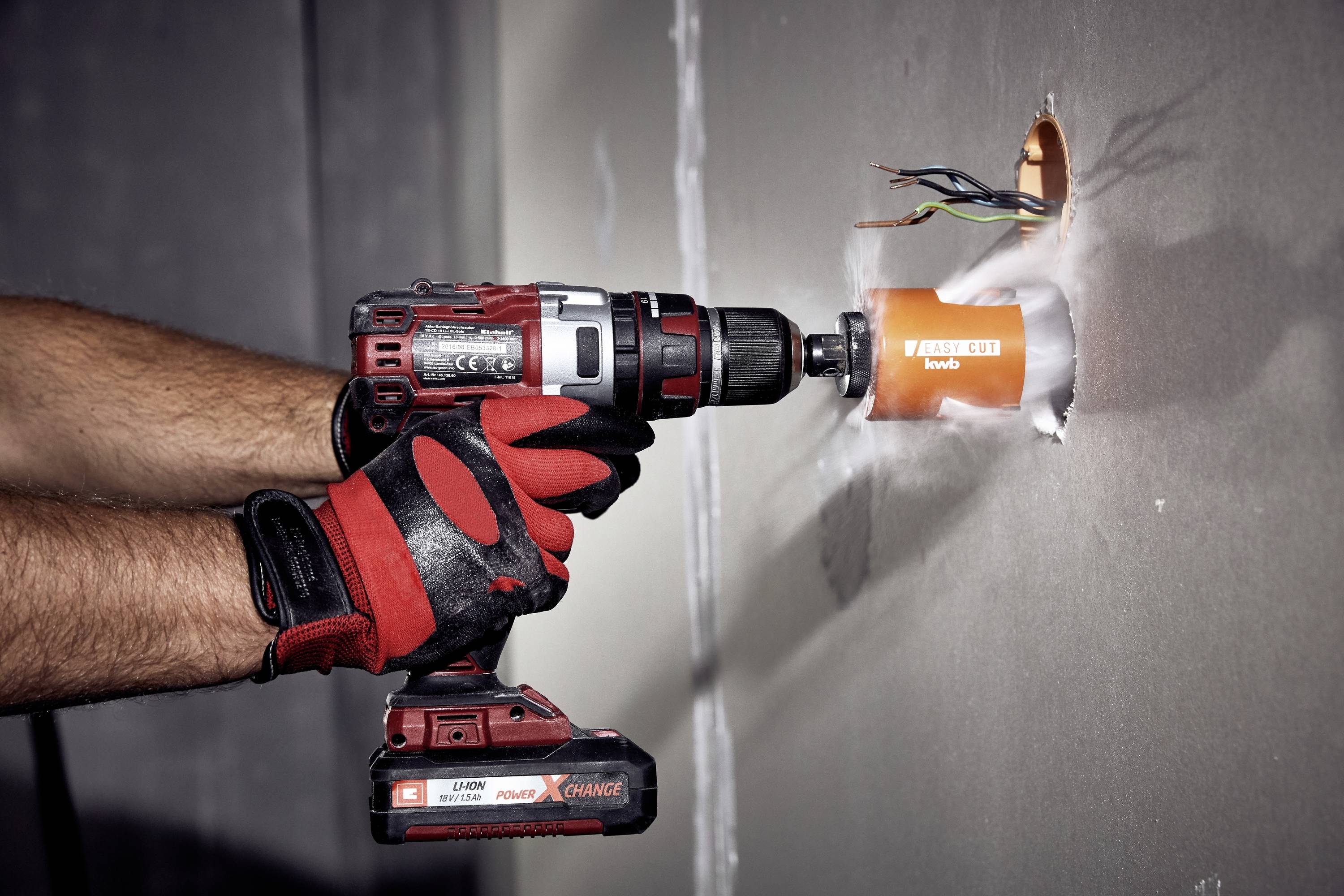 A person is drilling a round hole in a plasterboard wall using a cordless drill. Cables are protruding from the hole.