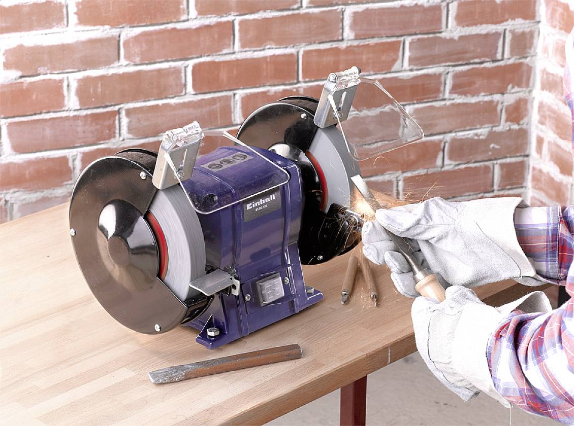 A person is grinding metal on a double-sided grinding wheel, creating sparks. The grinding wheel is positioned on a wooden table.