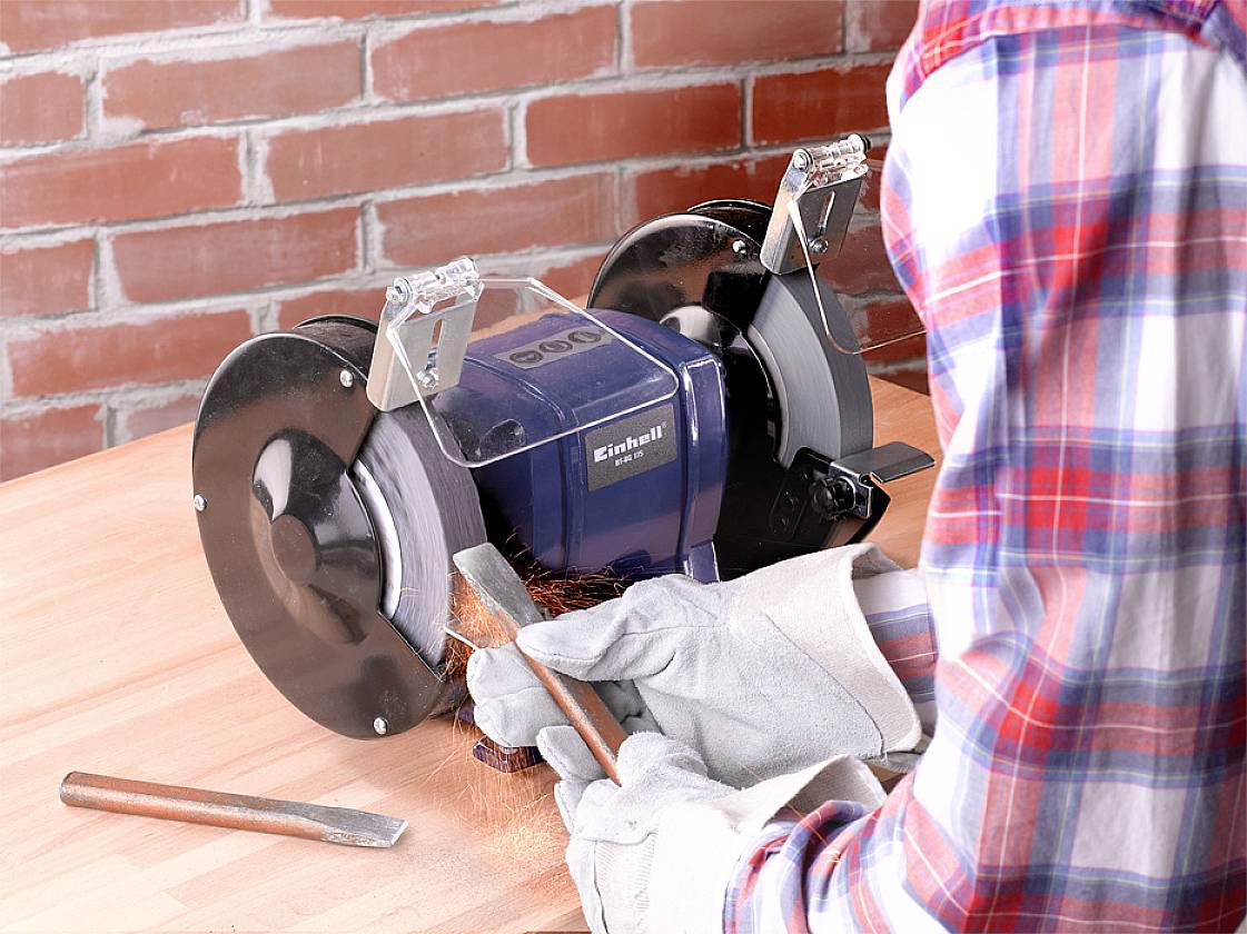 A person is grinding a metal tool with a grinding device, wearing gloves. Sparks are flying around the tool.