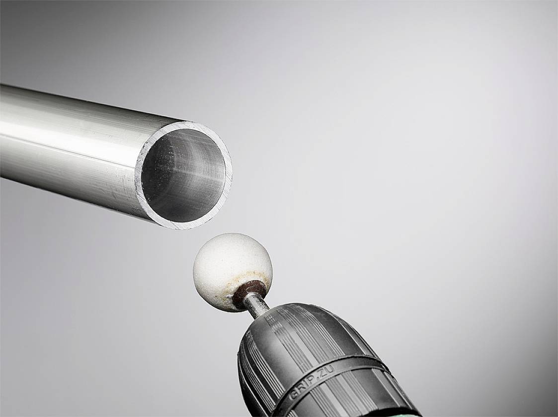 An aluminium tube is polished by a drill with a spherical nozzle. The background is grey and plain.