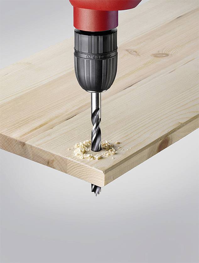A red electric drill is making a hole in a wooden board. Wood shavings are visible around the drill hole.