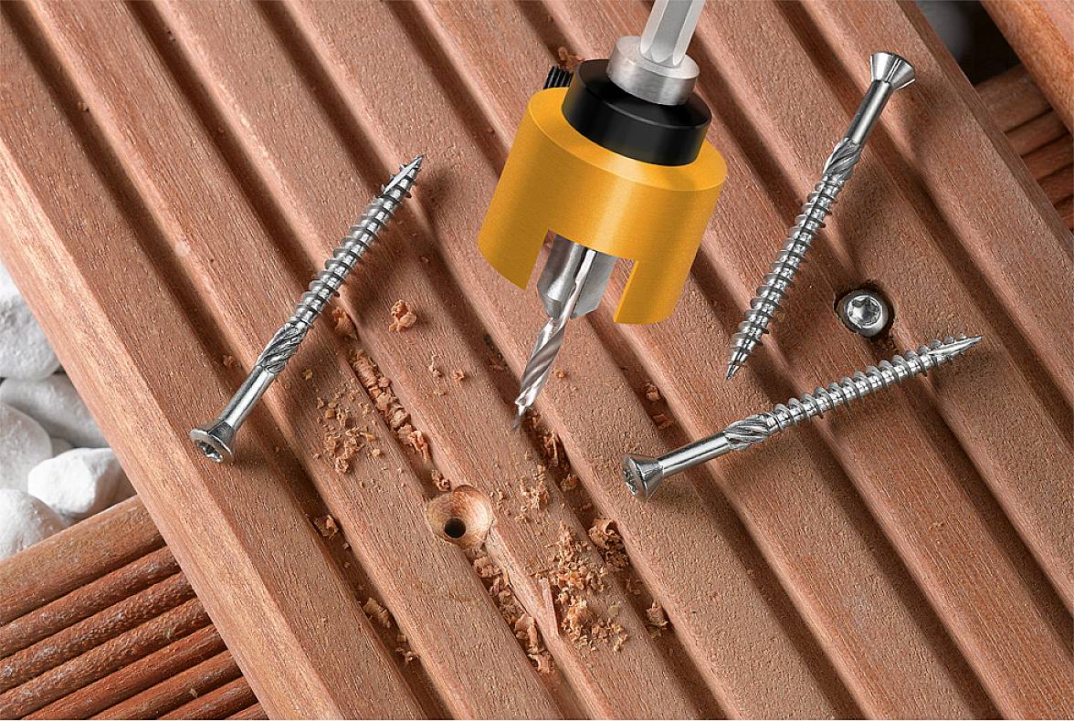 Wooden terrace with four screws, a yellow-orange drill bit, and a drilled hole in the centre, surrounded by wood shavings.