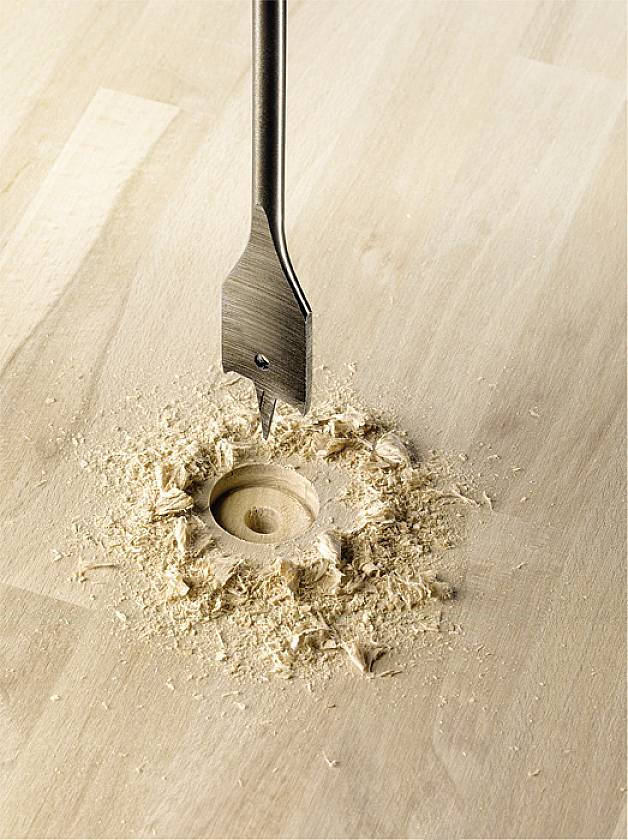 A flat drill bit is drilling a round hole in a wooden board, surrounded by wood shavings.